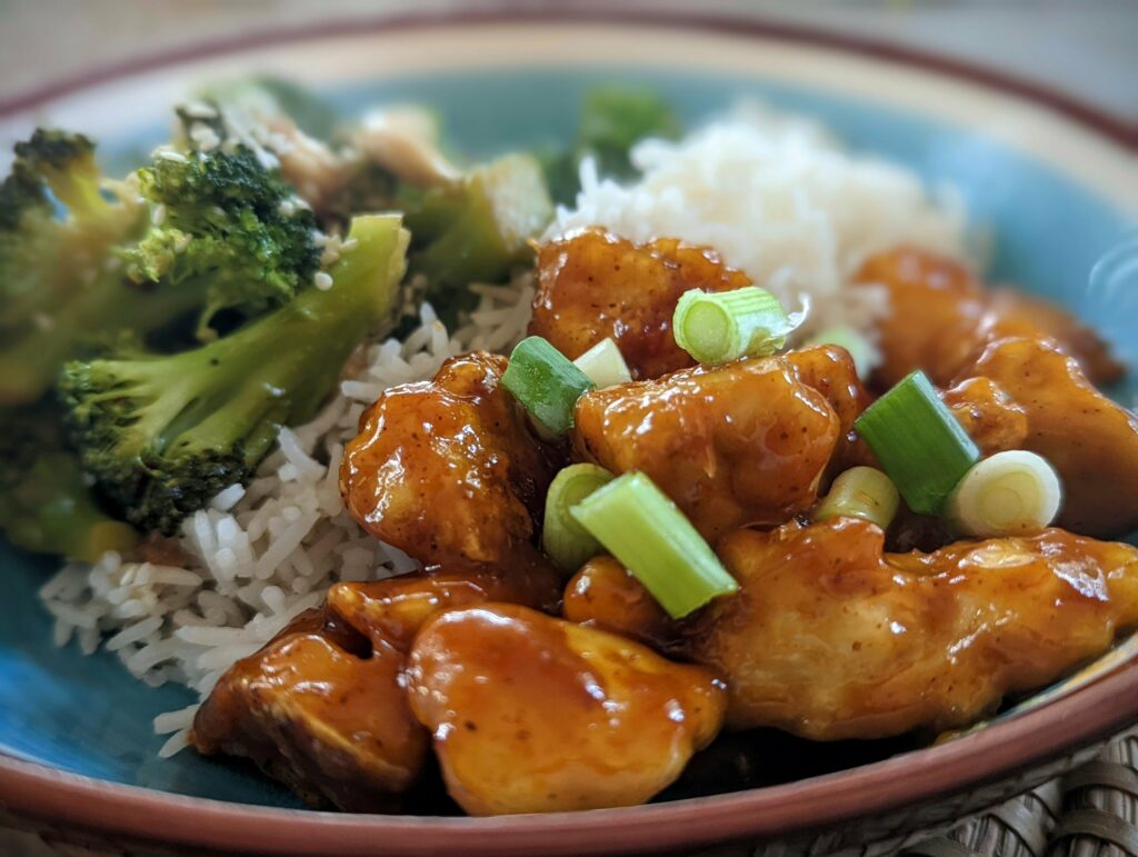 Pieces of glazed vegan chicken topped with chopped scallions, served with white rice and steamed broccoli in a blue-glazed bowl.