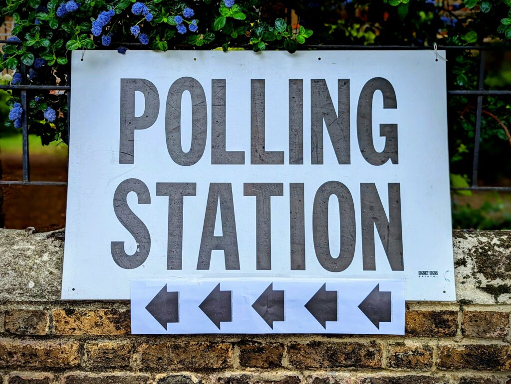 A white POLLING STATION sign with five black arrows pointing left, mounted on a fence above a brick wall with blue flowers behind it.