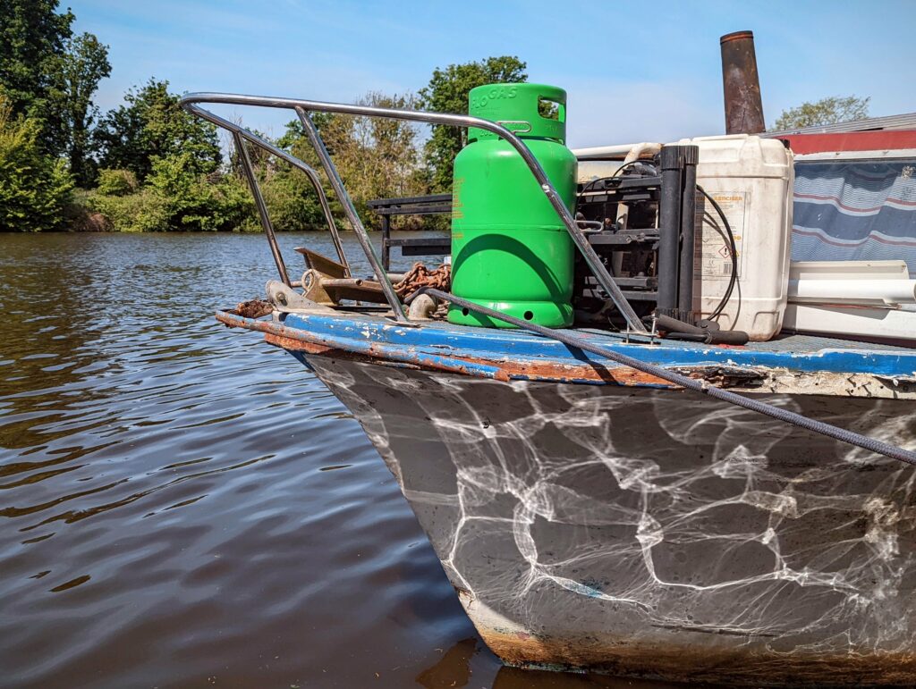 Sunlight reflects in shimmering patterns on the hull of a boat bow carrying a green gas cylinder on a tree-lined river.