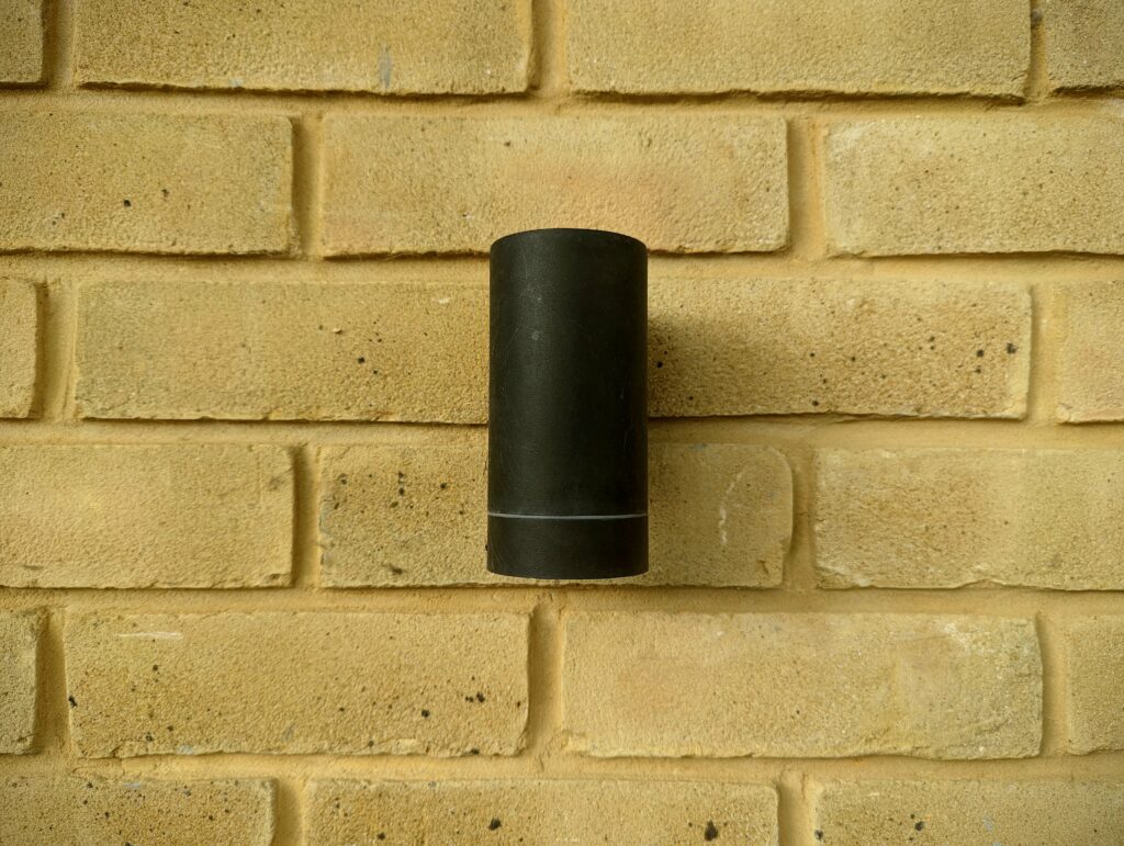 A black matte cylindrical wall light mounted vertically on a pale yellow brick wall.