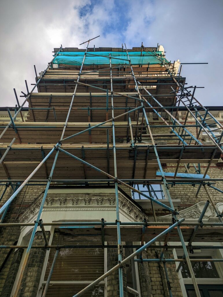 Low-angle view looking up at an ornate brick building encased in metal scaffolding and wooden platforms against a bright, cloudy sky.