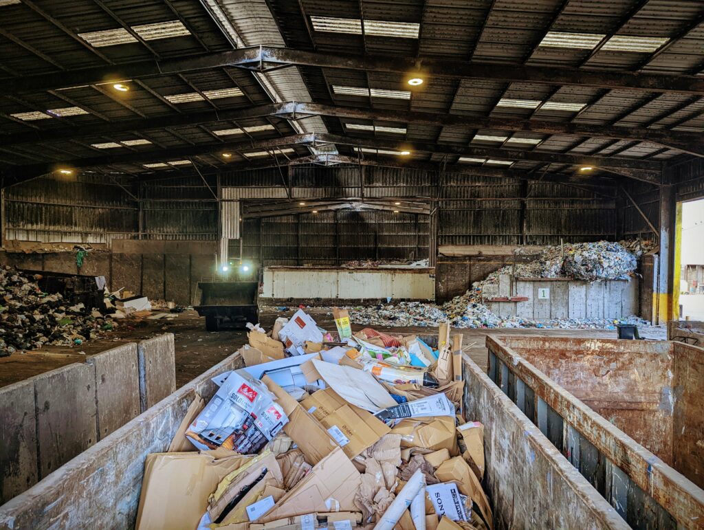 Cardboard recycling in metal skips inside a large waste management facility, with a front loader moving piles of trash in the background.