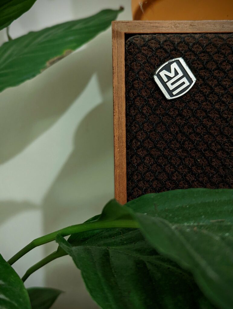 A vintage speaker with a brown mesh grille and a silver MS badge, framed by large green leaves of a houseplant.