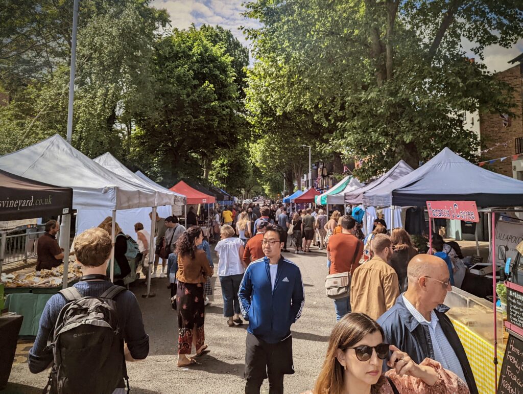 Crowds browsing a tree-lined street market with stalls under white and blue gazebos on a sunny day.