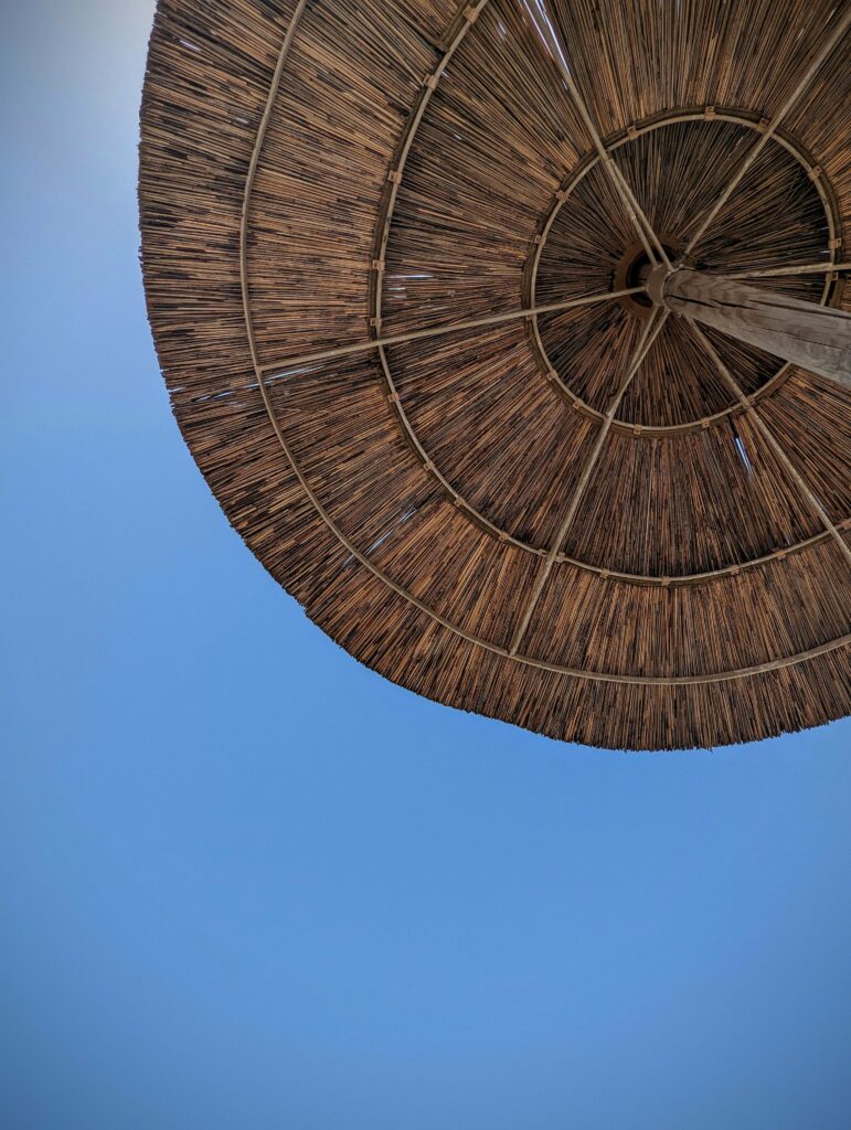 The underside of a circular thatched beach umbrella showing its texture and wooden frame against a clear blue sky.