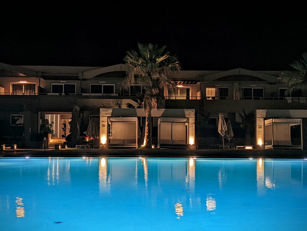 A glowing blue swimming pool at night reflecting warm lights from poolside cabanas and a central palm tree.