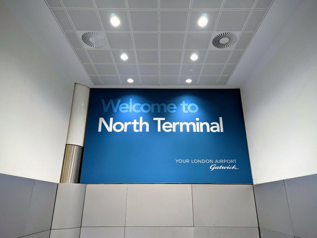 A blue sign at Gatwick Airport reading Welcome to North Terminal, Your London Airport Gatwick, under a grid of bright ceiling lights.
