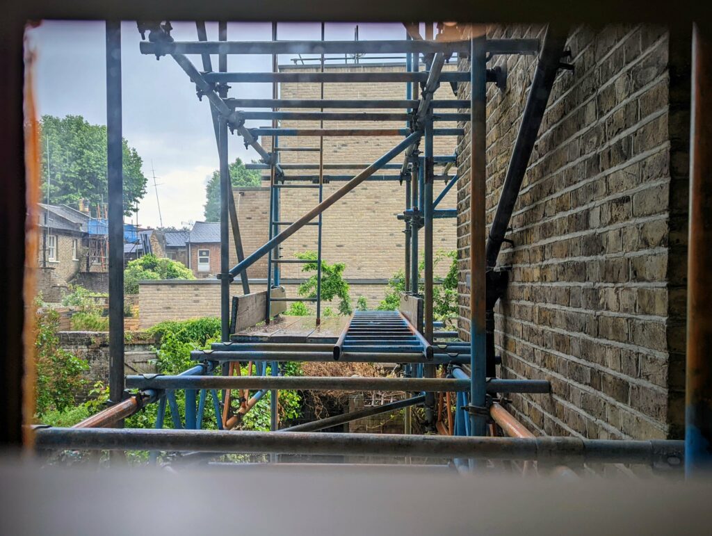 Blue scaffolding with wooden planks and a ladder erected against a brick wall, overlooking a garden and distant houses.