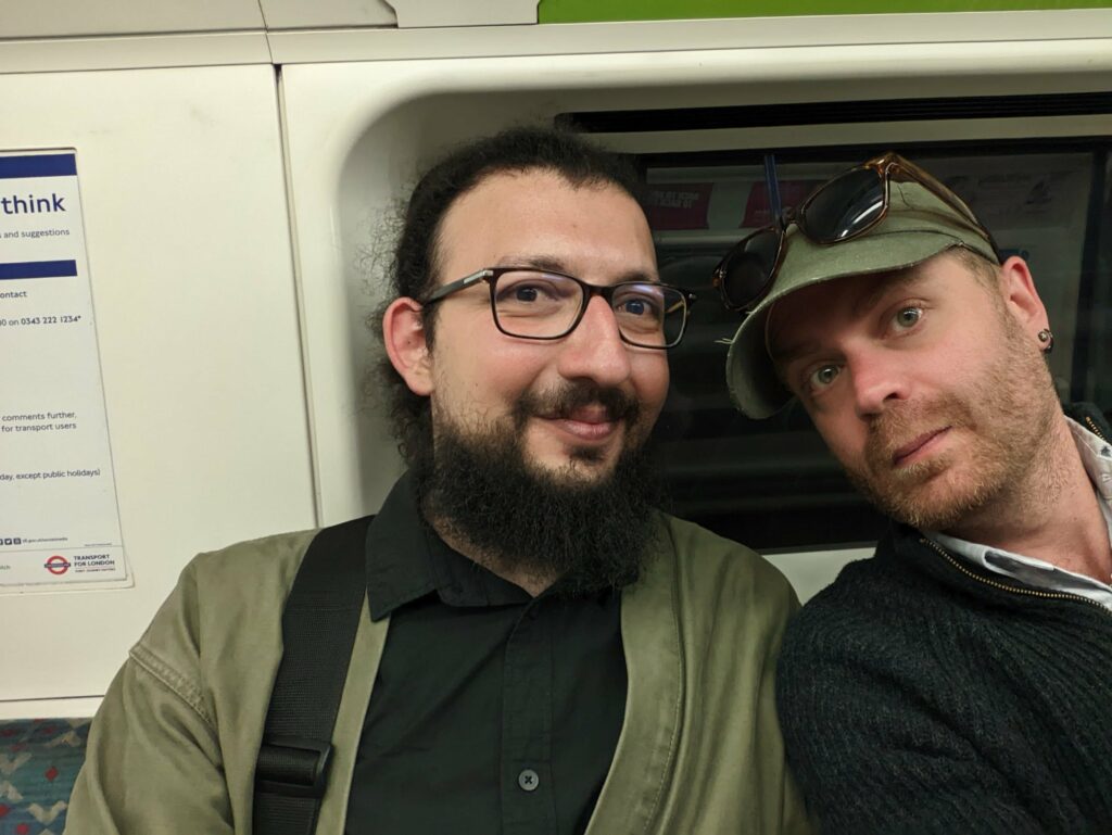 Ahmet Alphan Sabancı and Jay Springett taking a selfie on a London Underground train. One has a dark beard and glasses; the other wears a green cap with sunglasses.