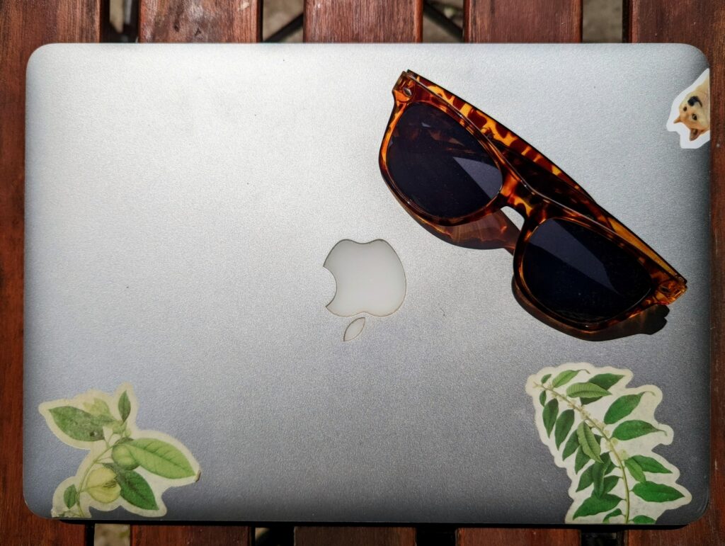 Tortoiseshell sunglasses on a silver MacBook lid with botanical stickers and a cat face, resting on a wooden slatted surface.