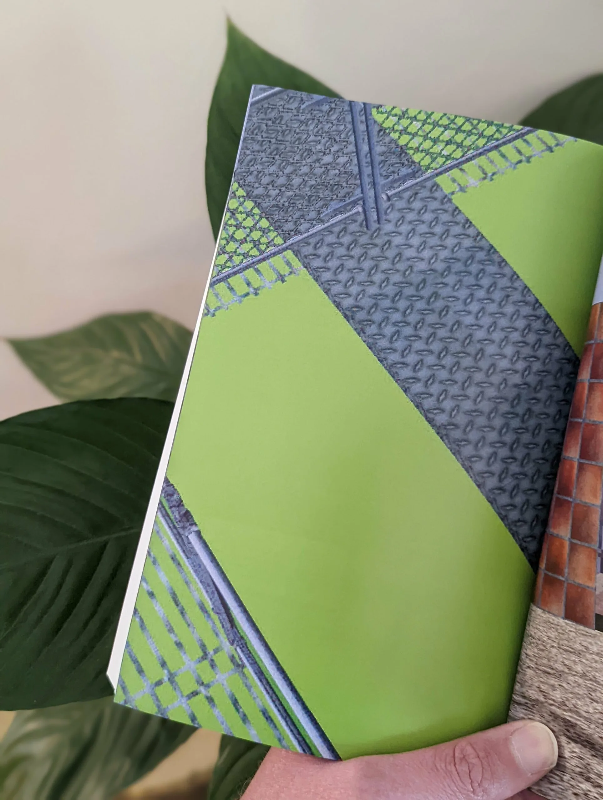 Lime green and grey diamond plate abstract patterns on a book page held against a backdrop of large green plant leaves.