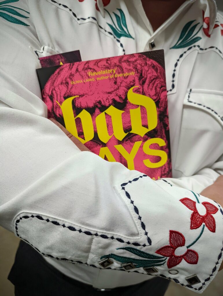 Huw Lemmey in a white western shirt with floral embroidery cradling the book Bad Gays, which features a bright pink and yellow cover.