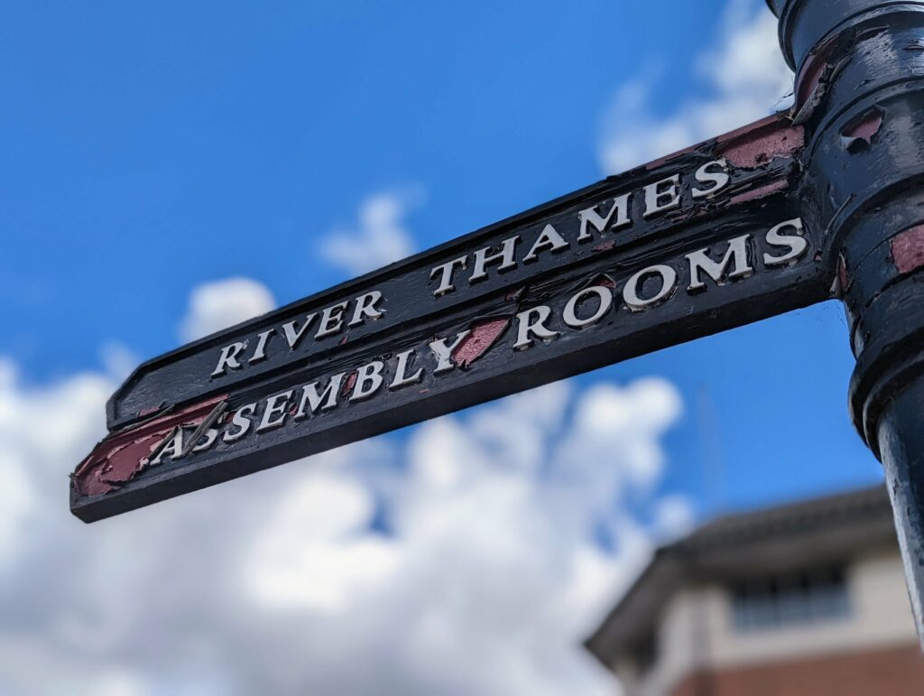 Weathered black fingerpost sign pointing to River Thames and Assembly Rooms against a bright blue sky with fluffy white clouds.