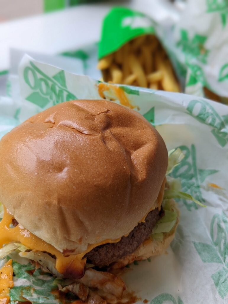 A cheeseburger with melted cheese and lettuce on green and white branded paper, with fries in the blurred background.