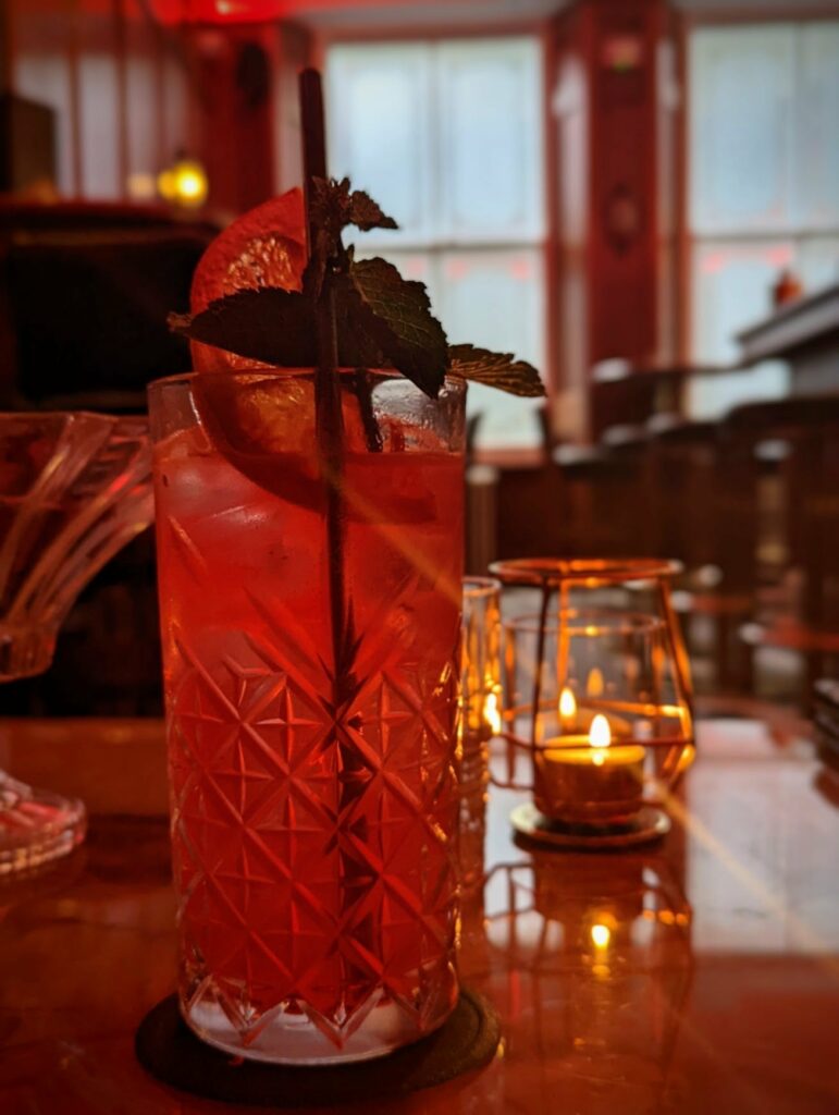 A red cocktail in a faceted glass garnished with blood orange and mint, set on a candlelit table in a dimly lit, atmospheric bar.