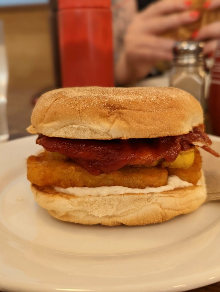 A breakfast roll filled with golden hash browns, bacon, red sauce, and white sauce on a white plate with a blurred cafe background.