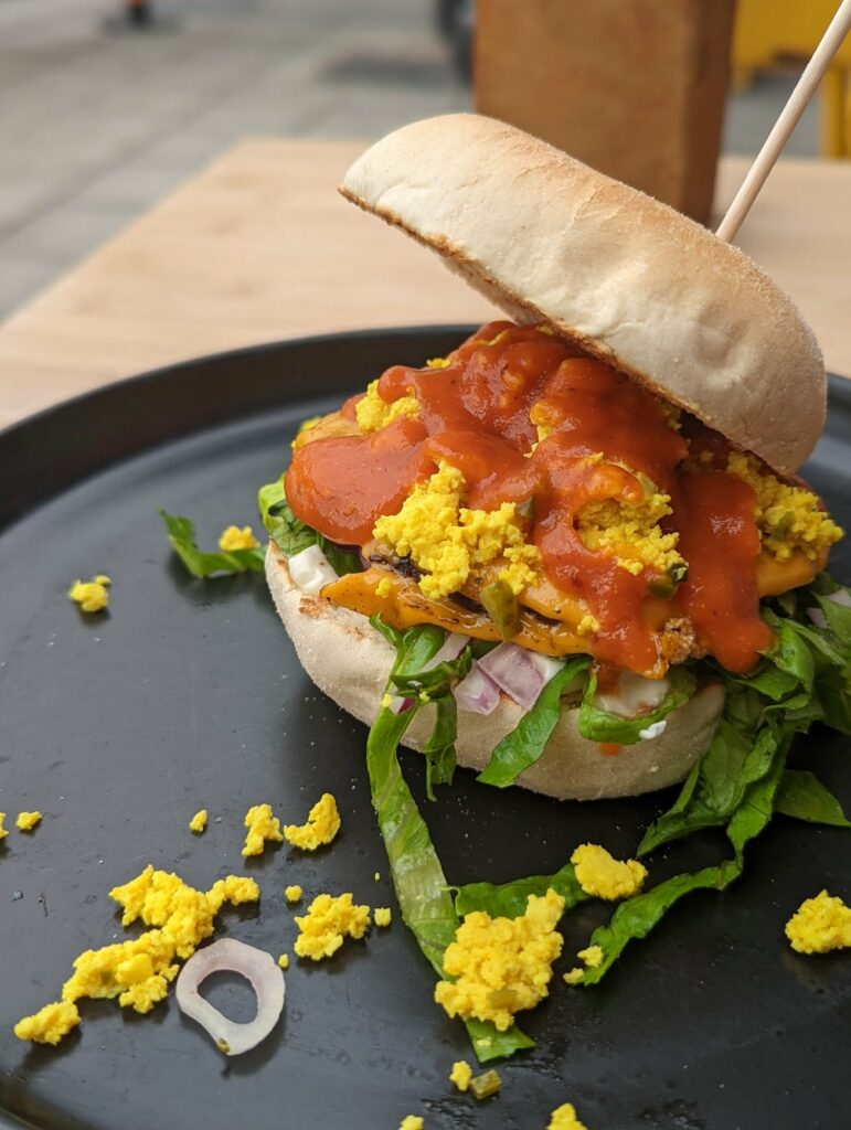 A tofu scramble breakfast burger topped with red sauce and lettuce in a toasted bun, served on a black plate.