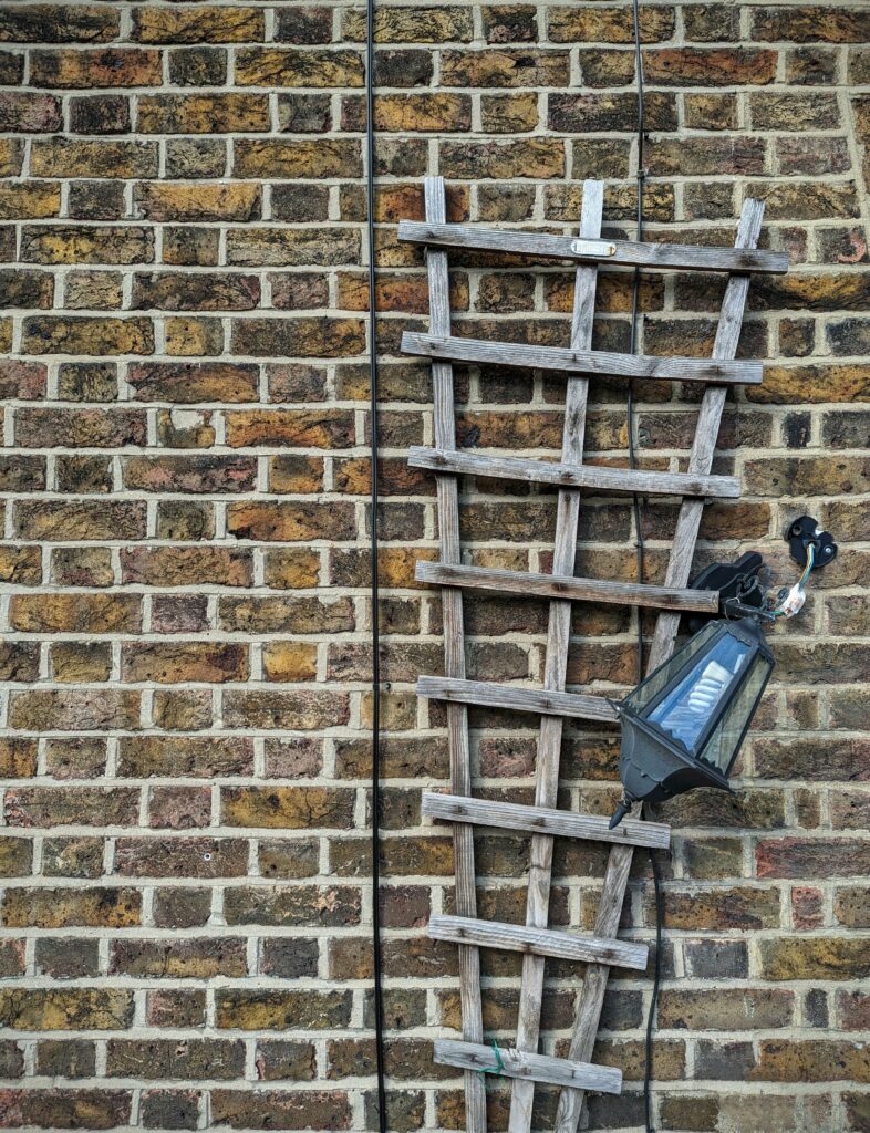 A weathered wooden garden trellis on a brick wall next to a black outdoor lamp hanging by its wires.