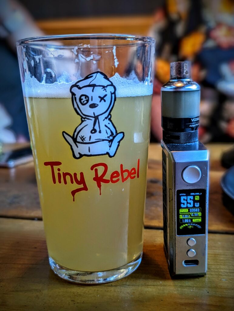Hazy beer in a Tiny Rebel glass with a teddy bear logo next to a silver vape mod showing 55W on a wooden table.