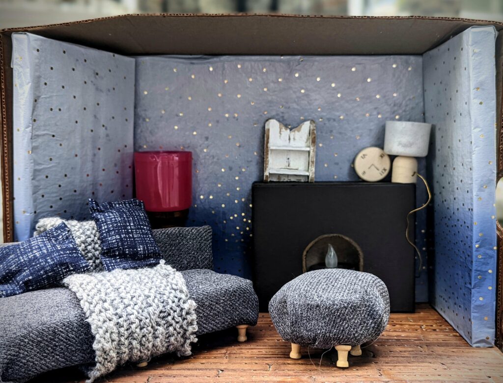 A miniature living room diorama with a grey sofa, chunky knit throw, ottoman, and black fireplace against blue wallpaper with gold dots.