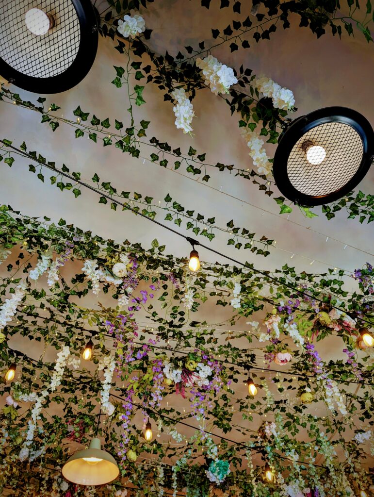 Ivy vines and floral garlands of white and purple flowers draped across a ceiling with industrial mesh lights and warm glowing bulbs.