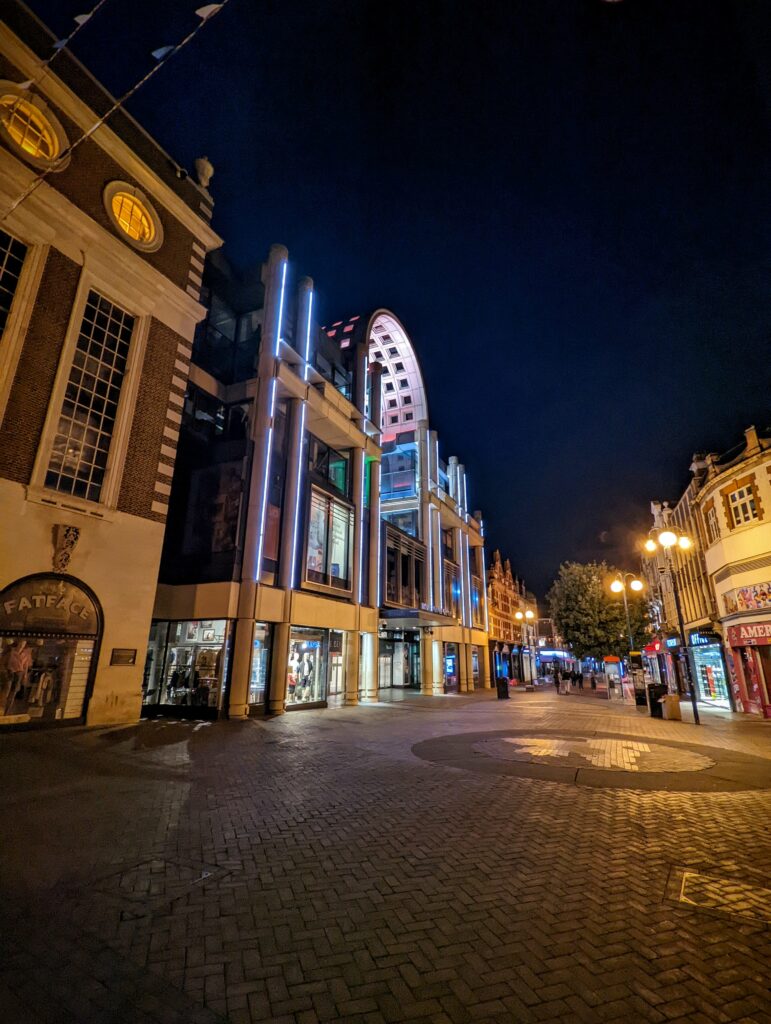 Illuminated shop fronts and a modern building with vertical blue lights and an arched glass roof on a cobbled pedestrian street at night.