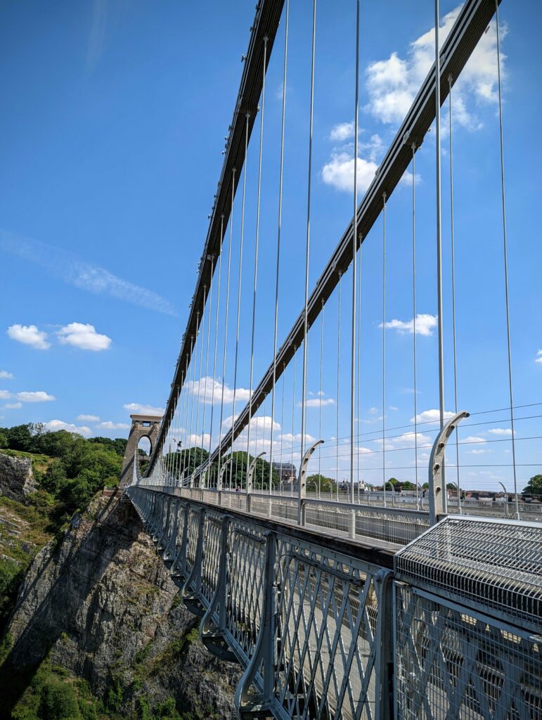 Cables and metal walkway of the Clifton Suspension Bridge stretching towards a stone tower against a bright blue sky with scattered clouds.