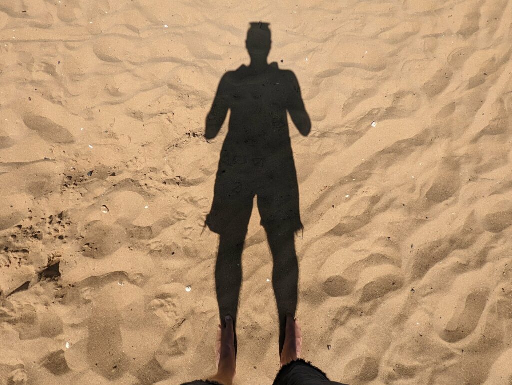 A person's long shadow cast across a sandy beach filled with footprints, with their feet visible at the bottom of the frame.