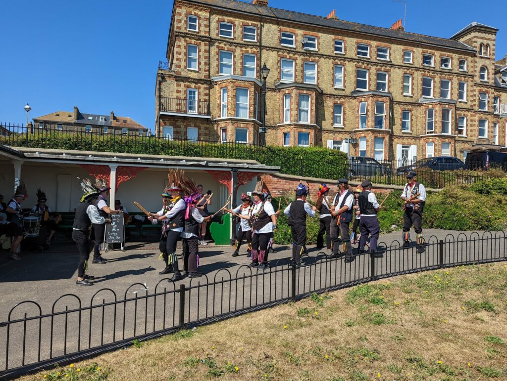 Morris dancers in feathered hats perform a stick dance in front of a seaside shelter in Broadstairs and grand yellow brick building on a sunny day.