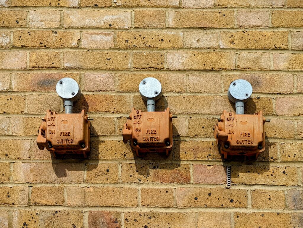Three weathered orange fire switch boxes mounted in a row on a yellow brick wall with silver metal conduits.