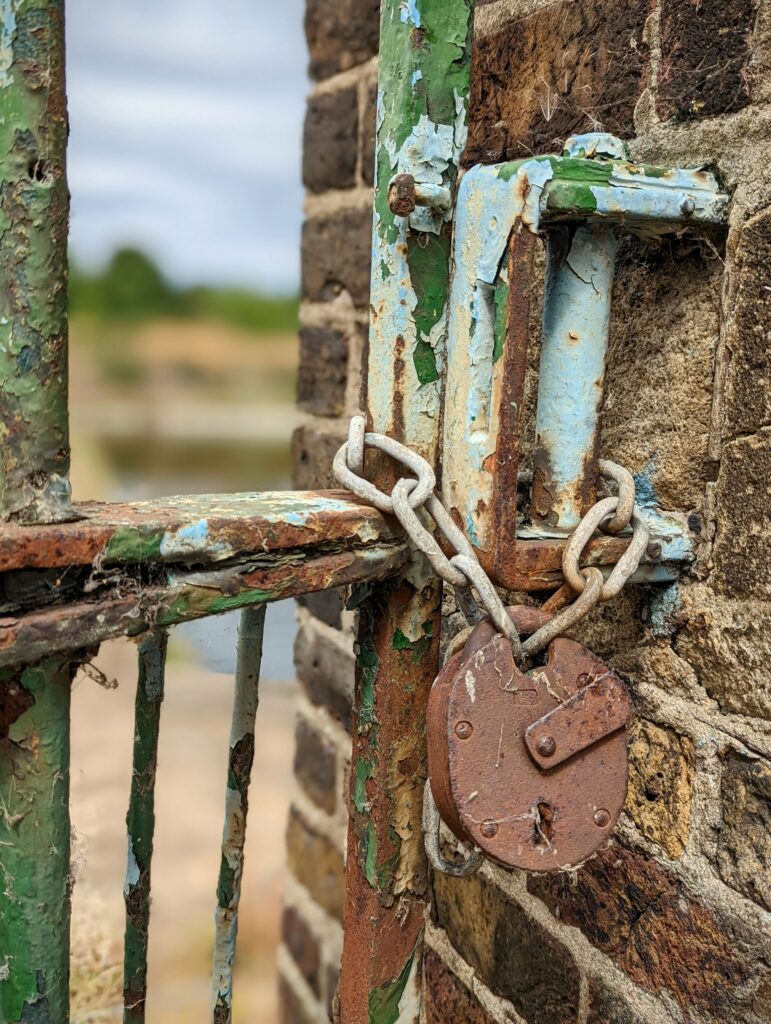 A rusty circular padlock and thick chain securing a weathered metal gate with peeling blue and green paint against an old brick wall.