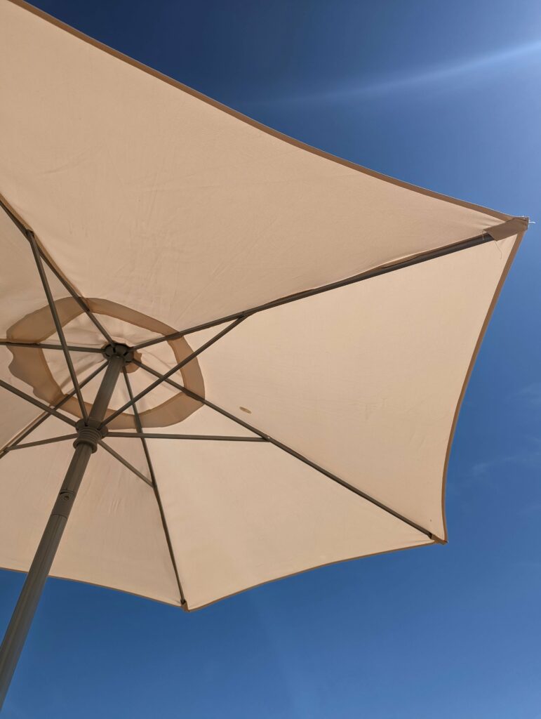 Looking up at the underside of a beige parasol showing its metal frame against a bright, cloudless blue sky.