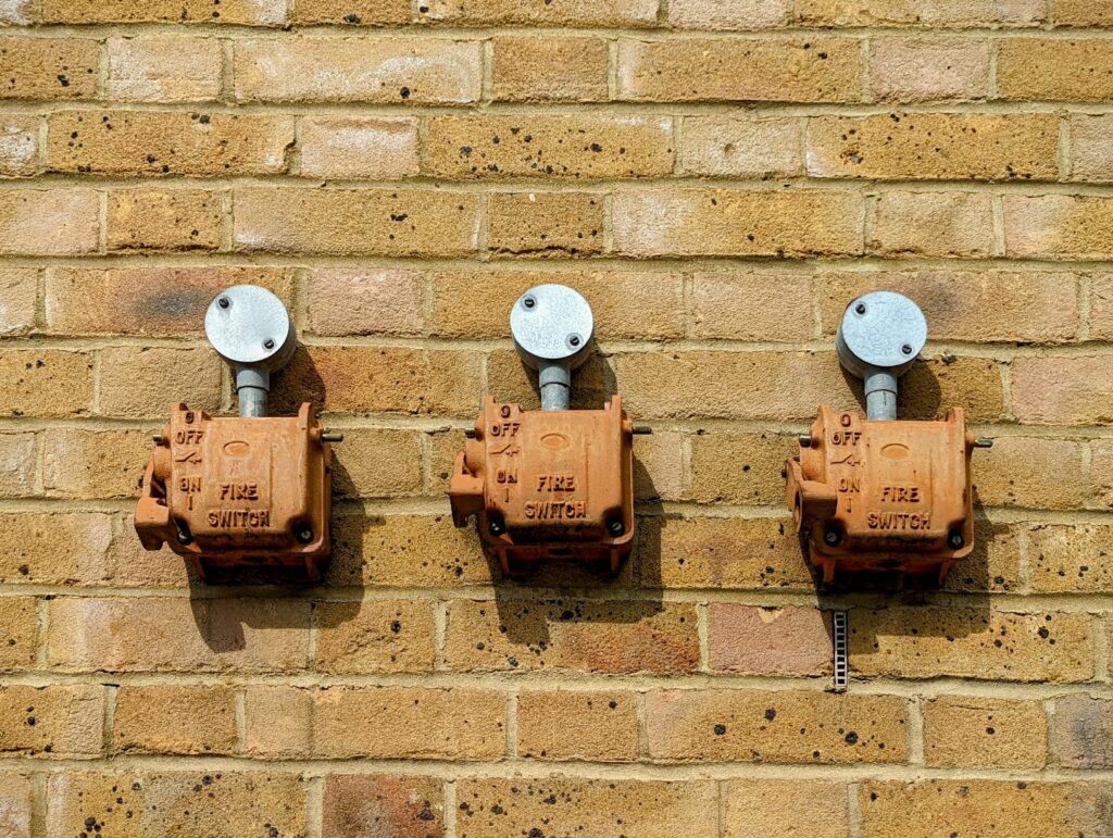 Three orange fire switches mounted in a row on a yellow brick wall with grey metal electrical conduits above each one.