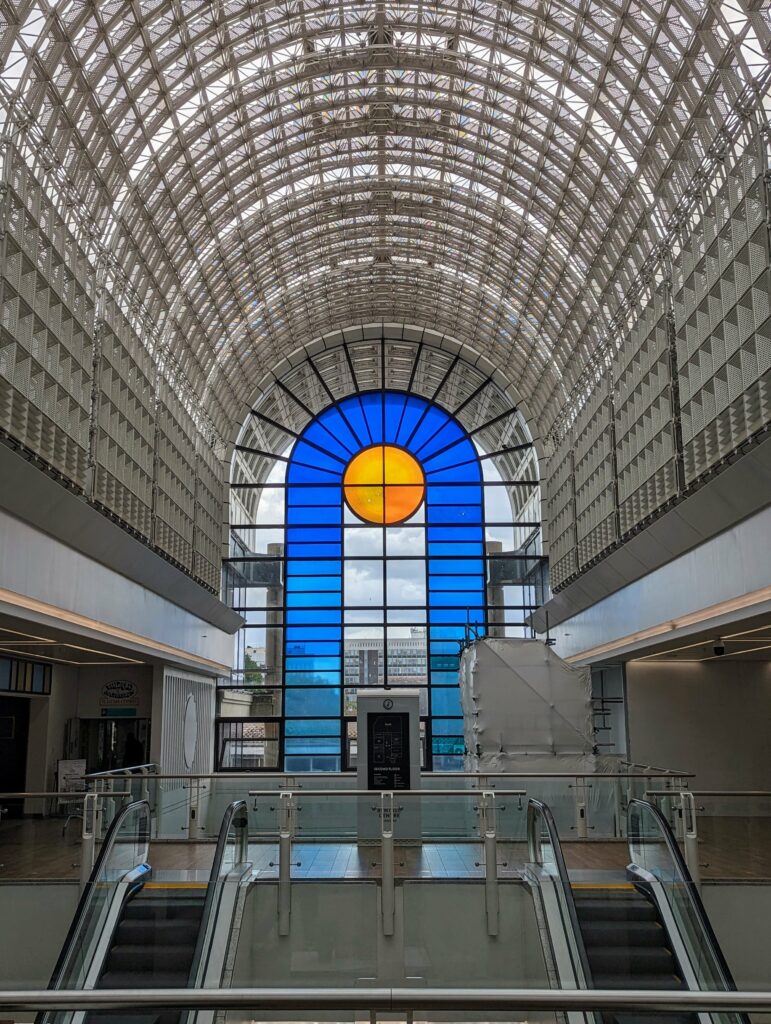 A vaulted lattice ceiling above a large arched window with blue panes and a central orange sun motif above two escalators.