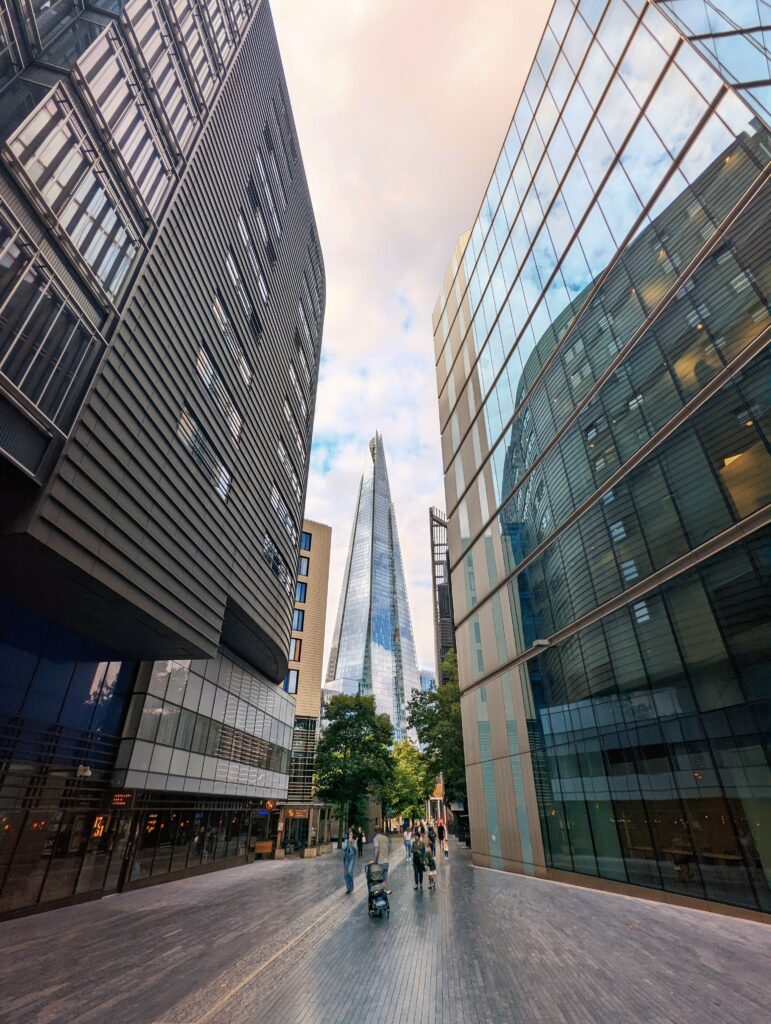 The Shard skyscraper framed by towering modern glass and metal buildings on a paved pedestrian walkway in London.