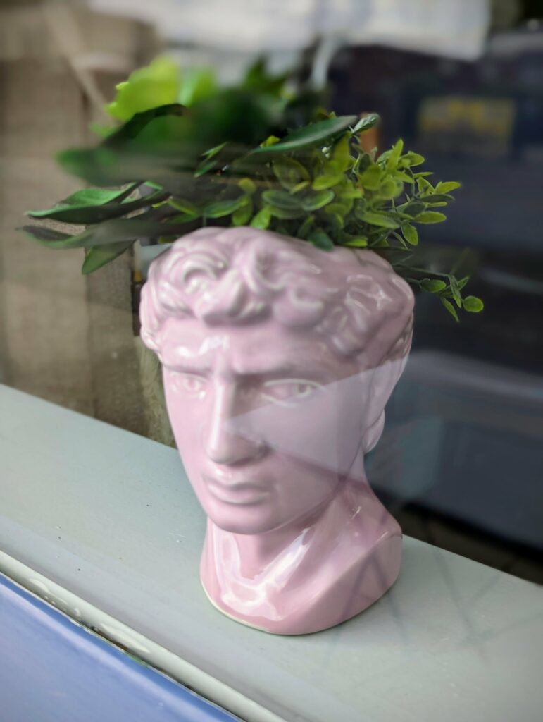 Pink glossy ceramic classical bust planter with green plants growing from the top, sitting on a white ledge.