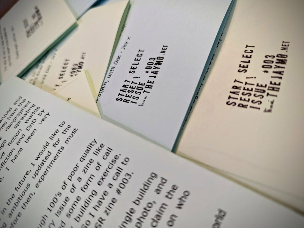 Stacks of zines titled START SELECT RESET! ISSUE #003 from THEJAYMO.NET with black typewriter-style text on pale paper.