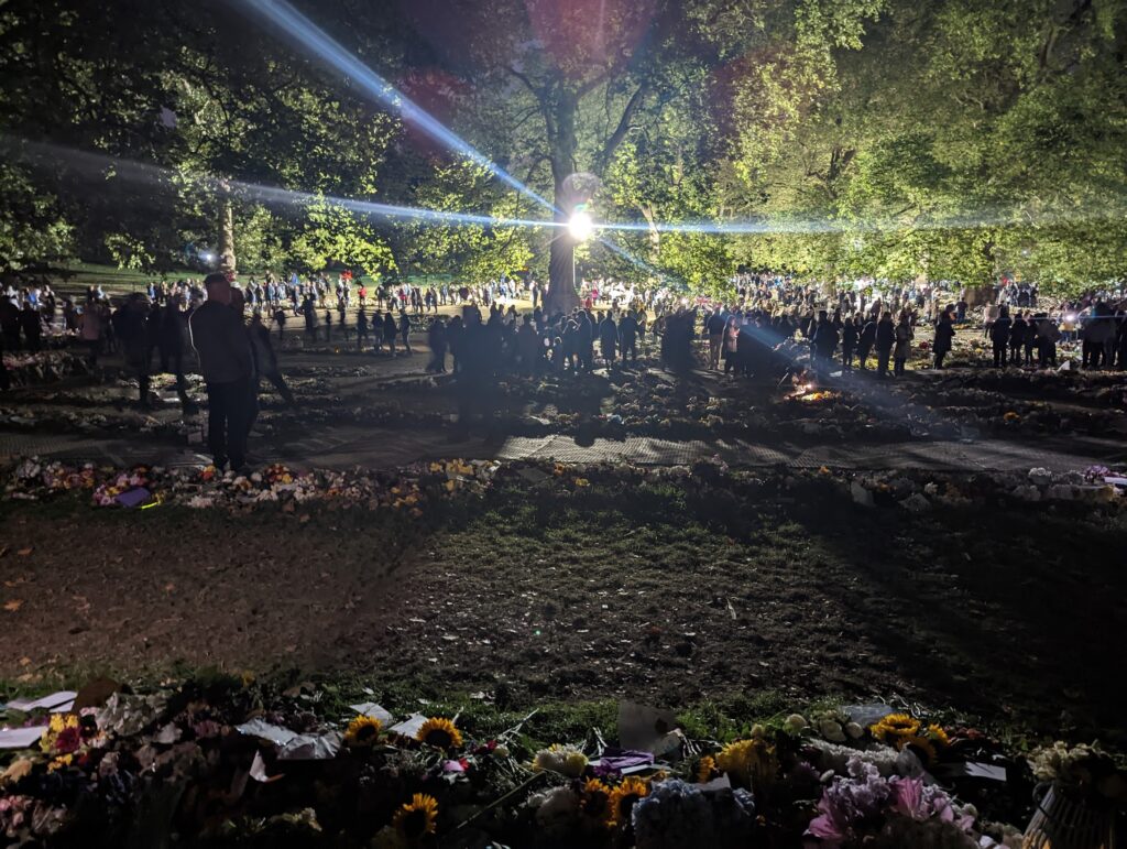 Crowds gathered at night among extensive rows of floral tributes in a park, with bright lens flares cutting across illuminated trees.