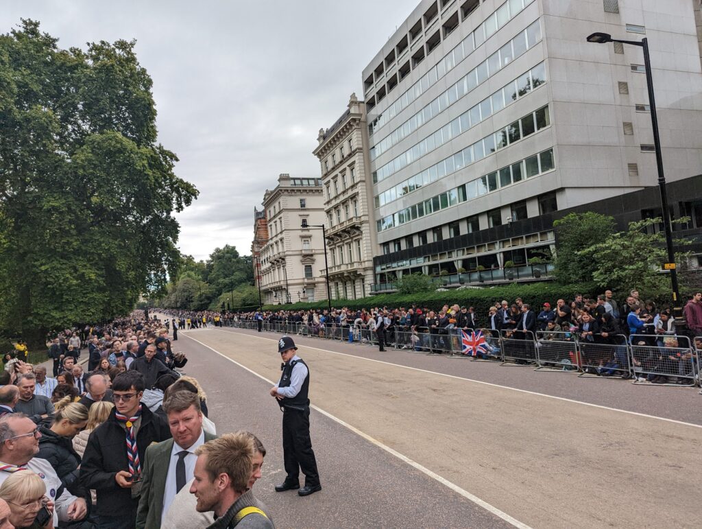 Crowds line metal barriers along a wide, empty London street under a grey sky, with a lone police officer standing in the road.