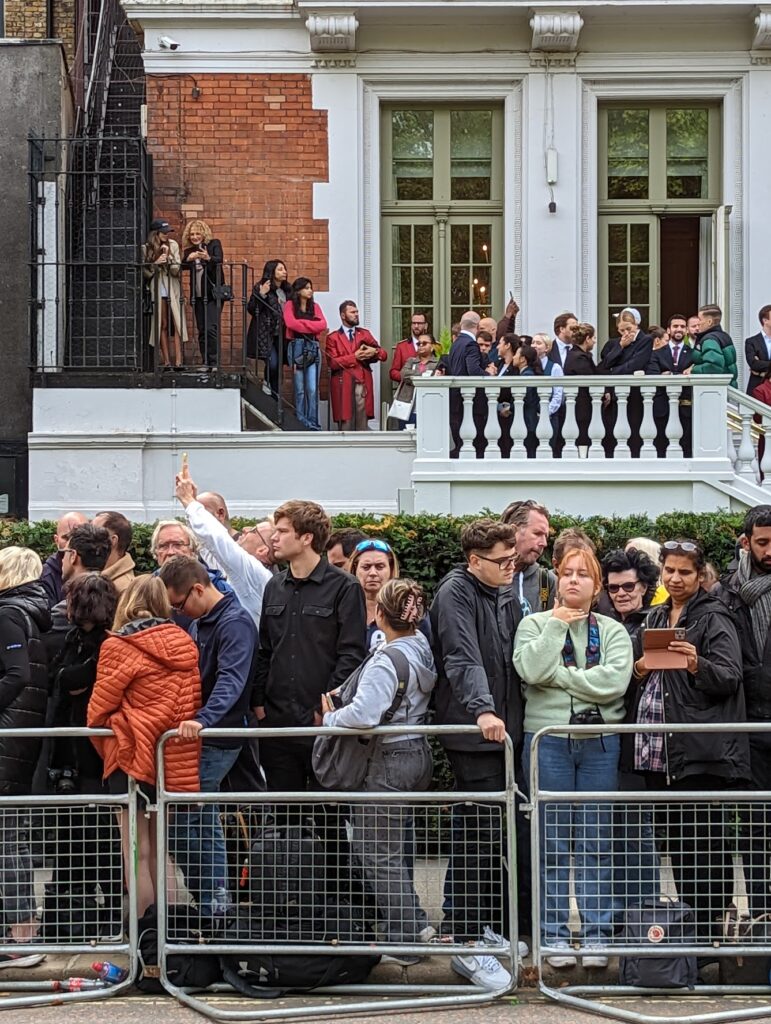 Crowds gathered behind metal barriers in London, with onlookers watching from the balcony and exterior stairs of a grand white building.