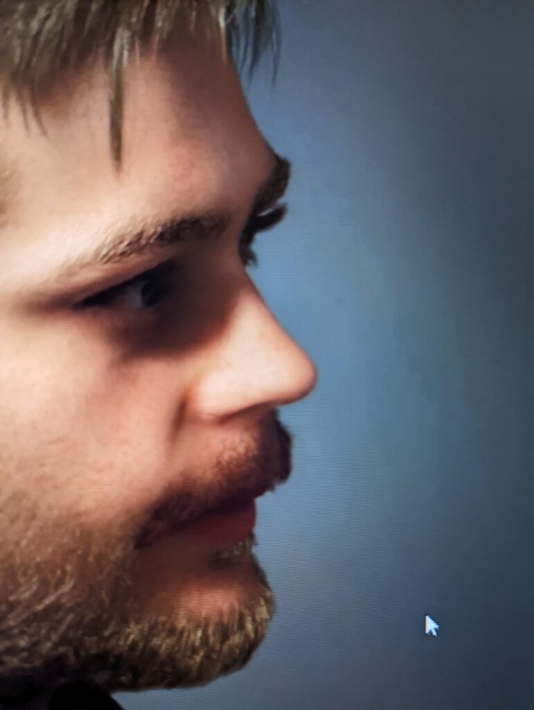 A close-up side profile of a man with fair hair and a short beard against a muted blue background.