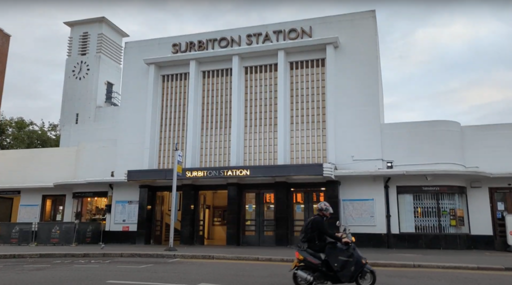 Art Deco facade of Surbiton Station with a white clock tower and bold signage, with a moped rider passing by on a grey day.