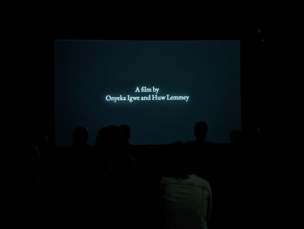 A projection screen in a dark room reading 'A film by Onyeka Igwe and Huw Lemmey' with silhouetted audience members in the foreground.