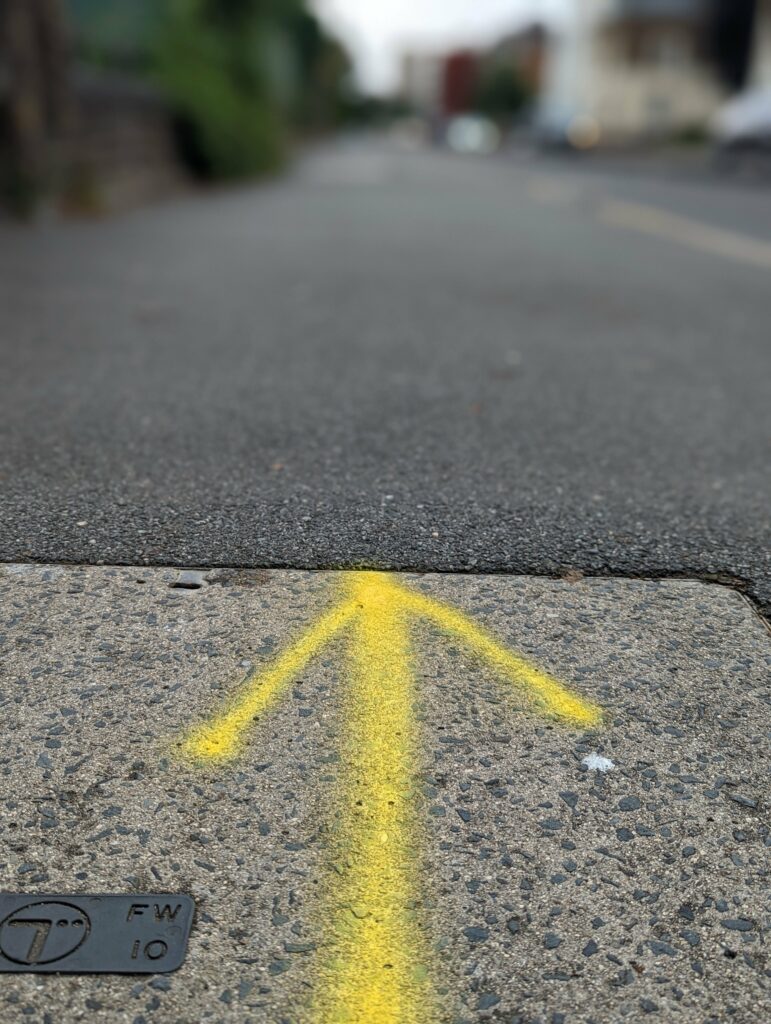 A yellow spray-painted arrow on a concrete pavement slab pointing toward a blurred asphalt road in a residential area.