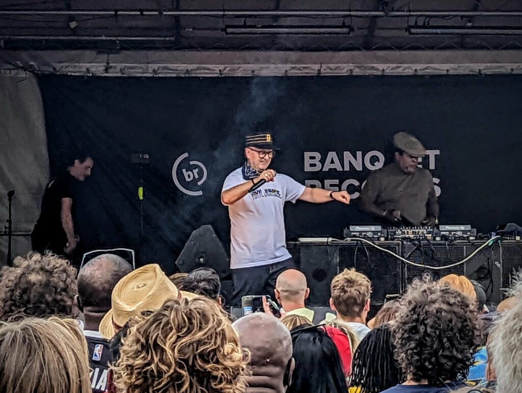 A performer in a white t-shirt and cap sings into a mic on an outdoor stage with a DJ behind him and a Banquet Records backdrop.