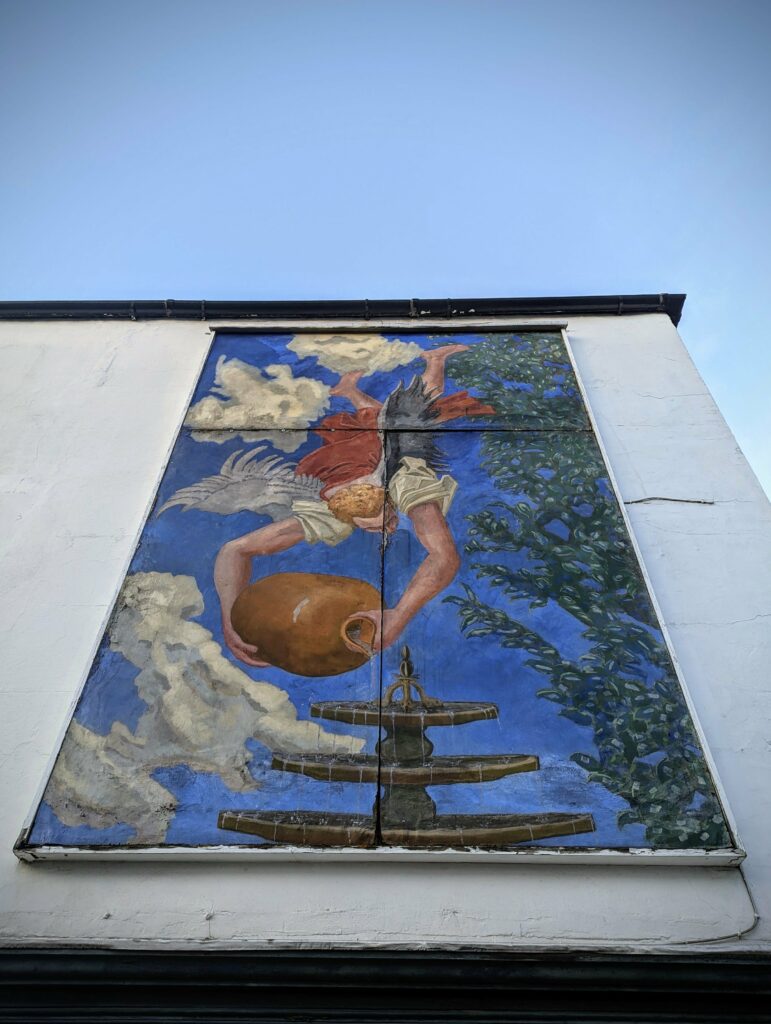 Mural on a white exterior wall depicting a winged figure pouring water from a large jug into a tiered fountain against a blue sky.