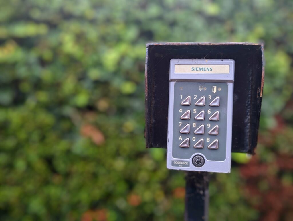 A grey Siemens Codelock security keypad mounted on a post in front of a blurred green garden hedge.