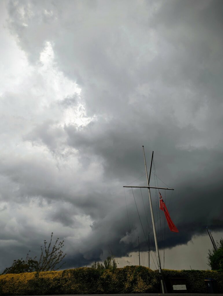 Dark, heavy storm clouds loom over a sailboat mast flying a Red Ensign flag above a yellow-green hedge.