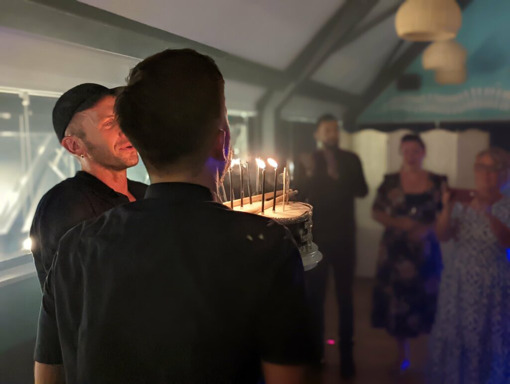 A man in a cap smiles at a lit birthday cake held by another person in a dimly lit room with guests in the blurred background.