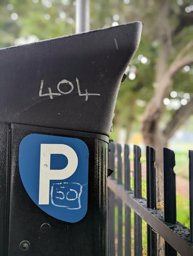 White ink graffiti reading 404 on a black parking meter. A blue P sticker features a boxed 50, set against a black fence and blurred trees.